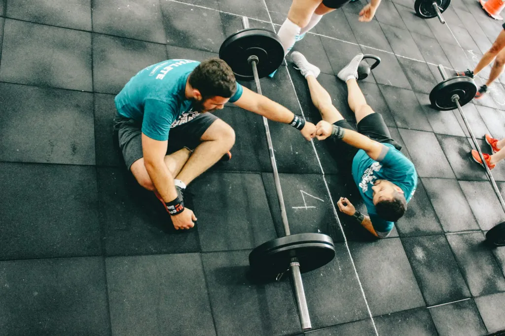Dos personas chocan los puños sentadas en el suelo de un gimnasio, junto a una barra con discos